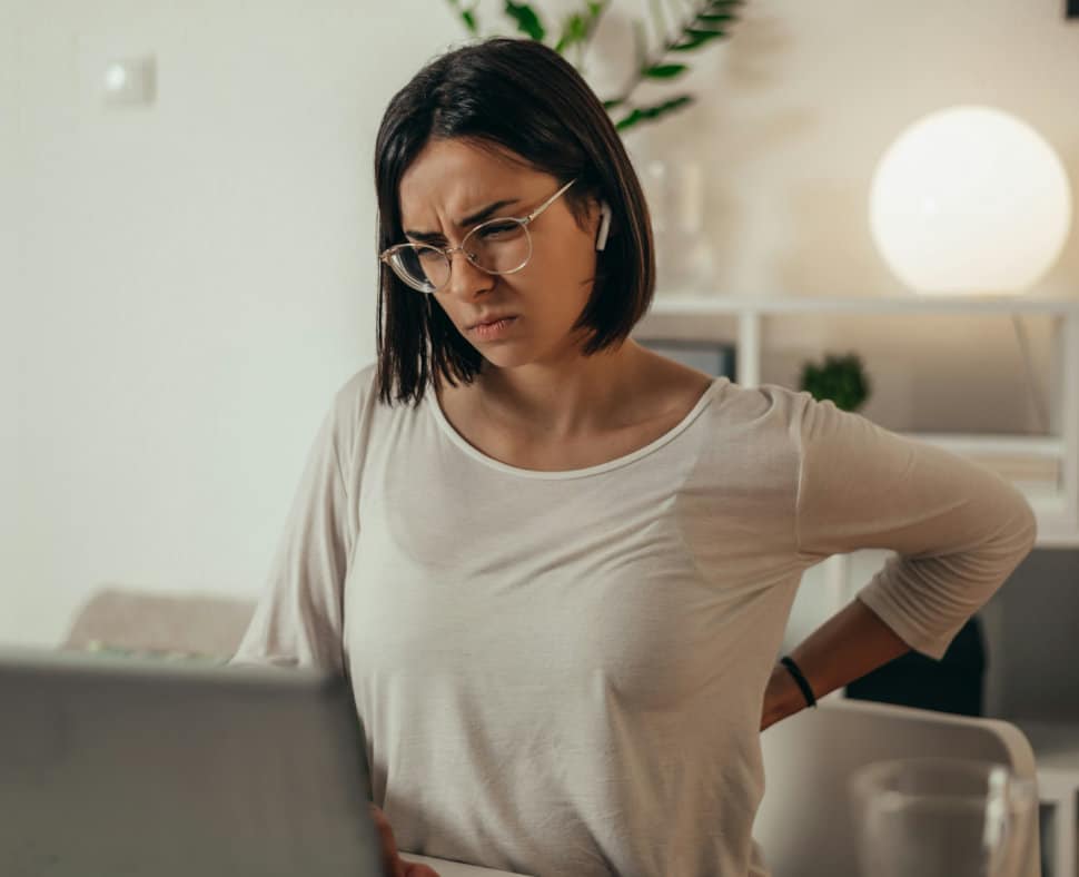 Une femme assise devant son ordinateur avec un mal de dos dû à sa mauvaise posture se tient le dos en grimaçant